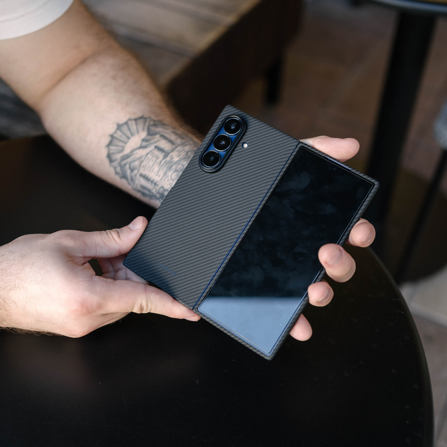 A person holds a black carbon-fiber-galaxy z fold 8 ultra protective case on a foldable smartphone, with the screen folded over. The case has a textured, woven-look finish and is shown in a real-world setting over a dark table.