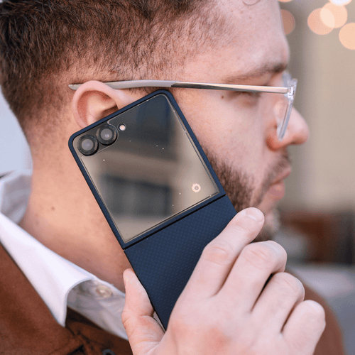A close-up lifestyle shot of a man holding a galaxy z flip 7 with a textured navy galaxy z flip 8 case cover against his ear, wearing thin metal-framed glasses.