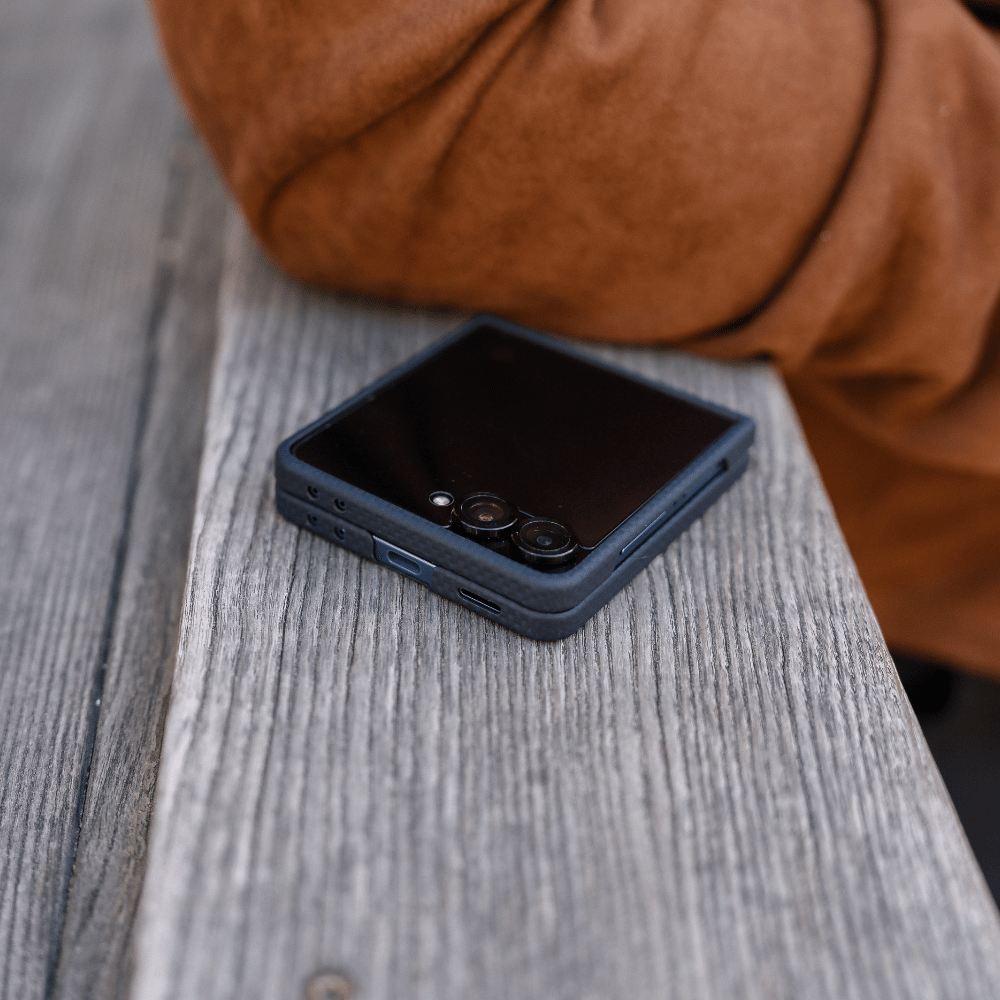 A navy galaxy z flip 7 case on a folded smartphone, resting on a weathered gray wooden surface with a person's brown sleeve visible in the background.