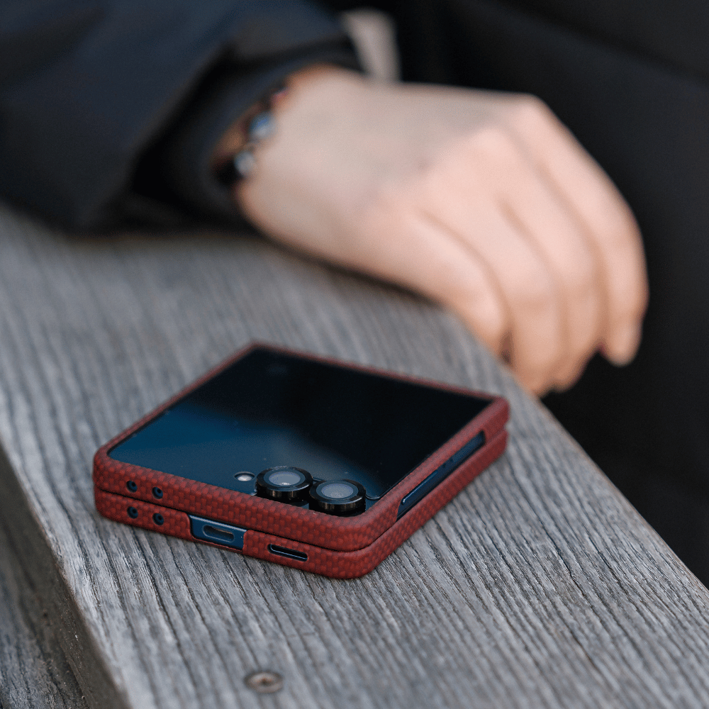 Close-up lifestyle shot of Samsung Galaxy Z Flip 8 with scarlet red aramid fiber phone case, placed on wooden table, detail view of charging port, speaker cutouts and camera lens frame, business daily carry scene with blurred hand background