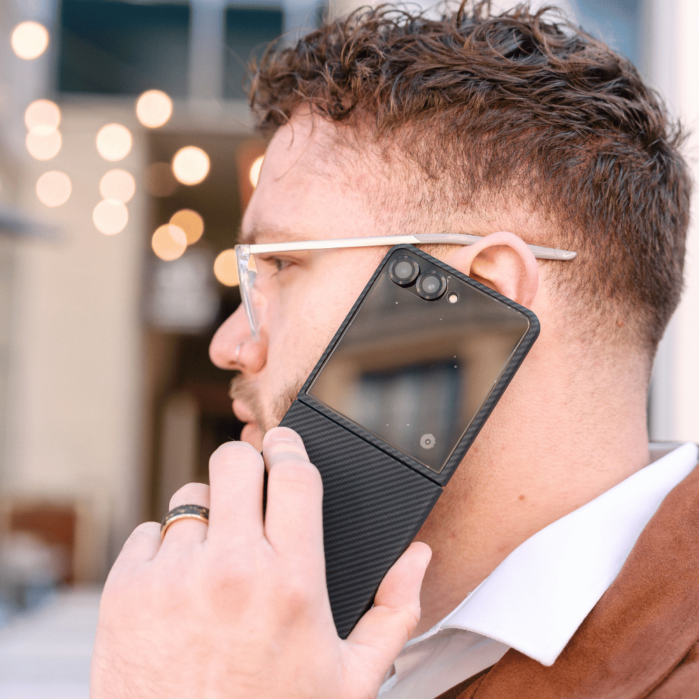 A man in profile wearing clear-framed glasses with a black magnetic galaxy z flip 7 case attached to his ear, holding a smartphone against his ear.