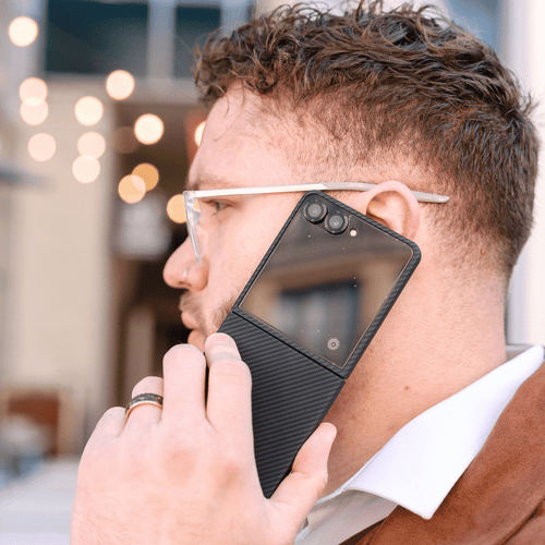 A man in profile wearing clear-framed glasses with a black magnetic galaxy z flip 7 case attached to his ear, holding a smartphone against his ear.
